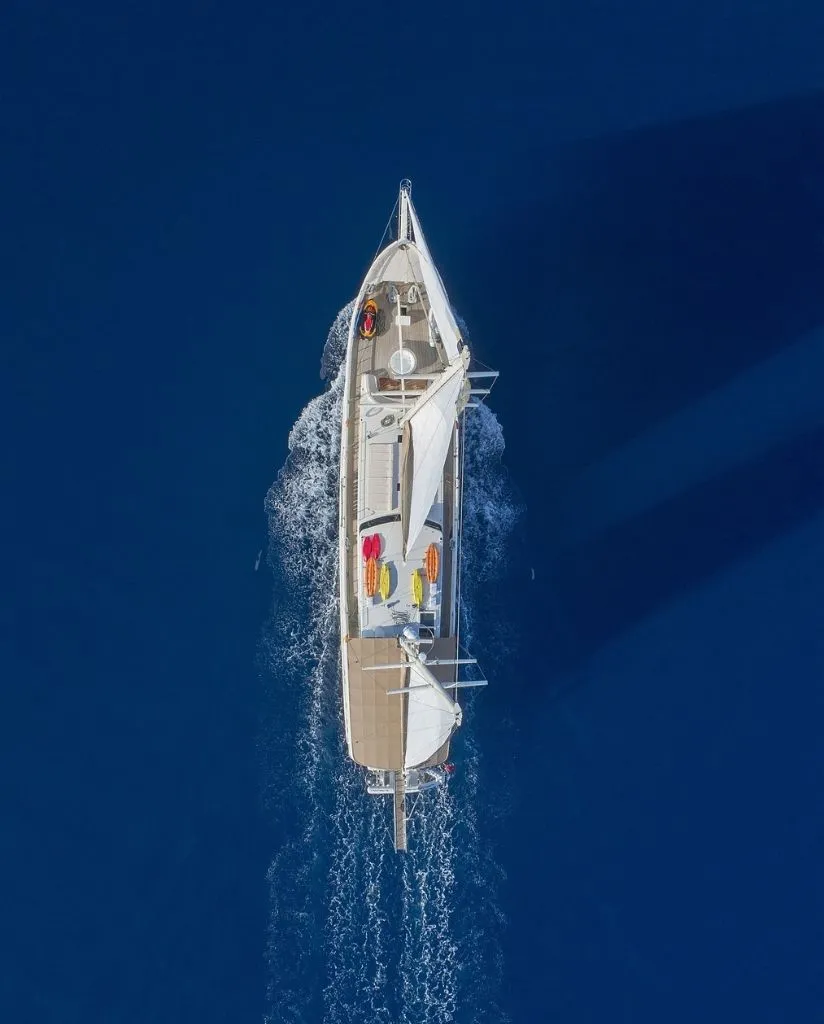 Aerial view of a white sailboat moving through deep blue water, leaving a trail of white waves behind it. The boat casts a large shadow on the water.