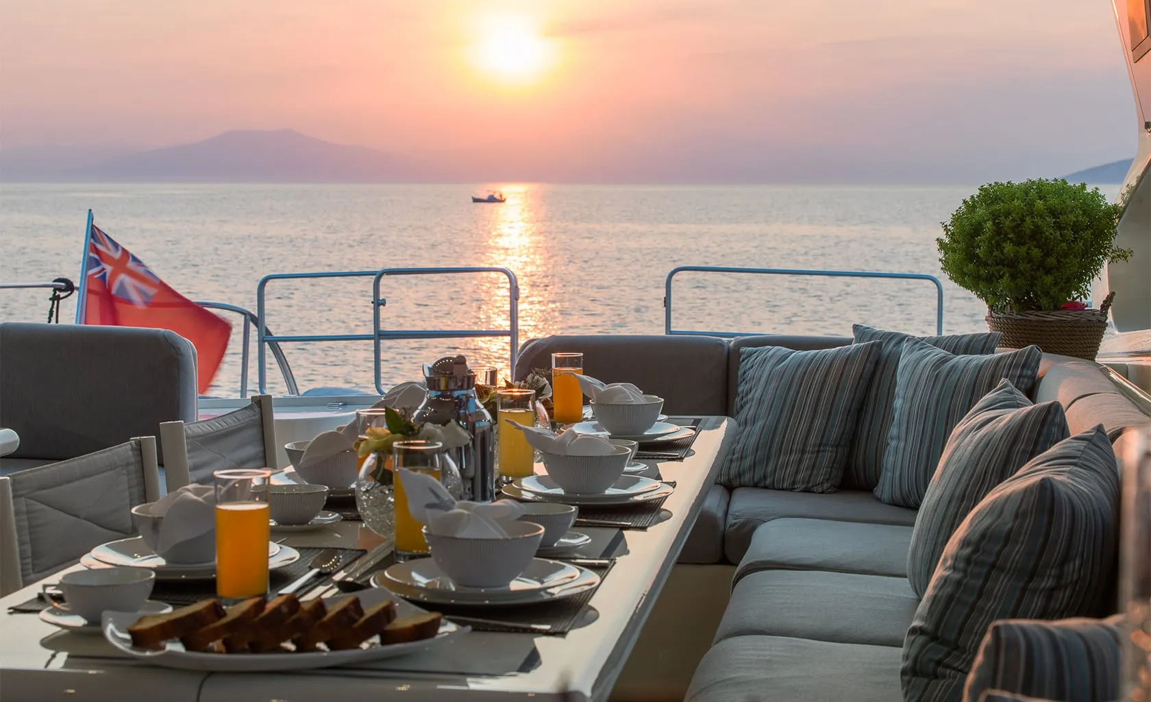 Breakfast table with dishes, glasses of juice, and toast set on a yacht deck with cushioned seating, overlooking a calm sea at sunset. A small boat is visible on the water and a mountain in the distance.