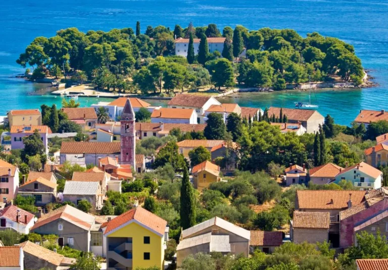 Colorful houses with red-tiled roofs cluster near a church tower, with lush greenery and a small, tree-covered island beyond, all beside bright blue water under a clear sky.