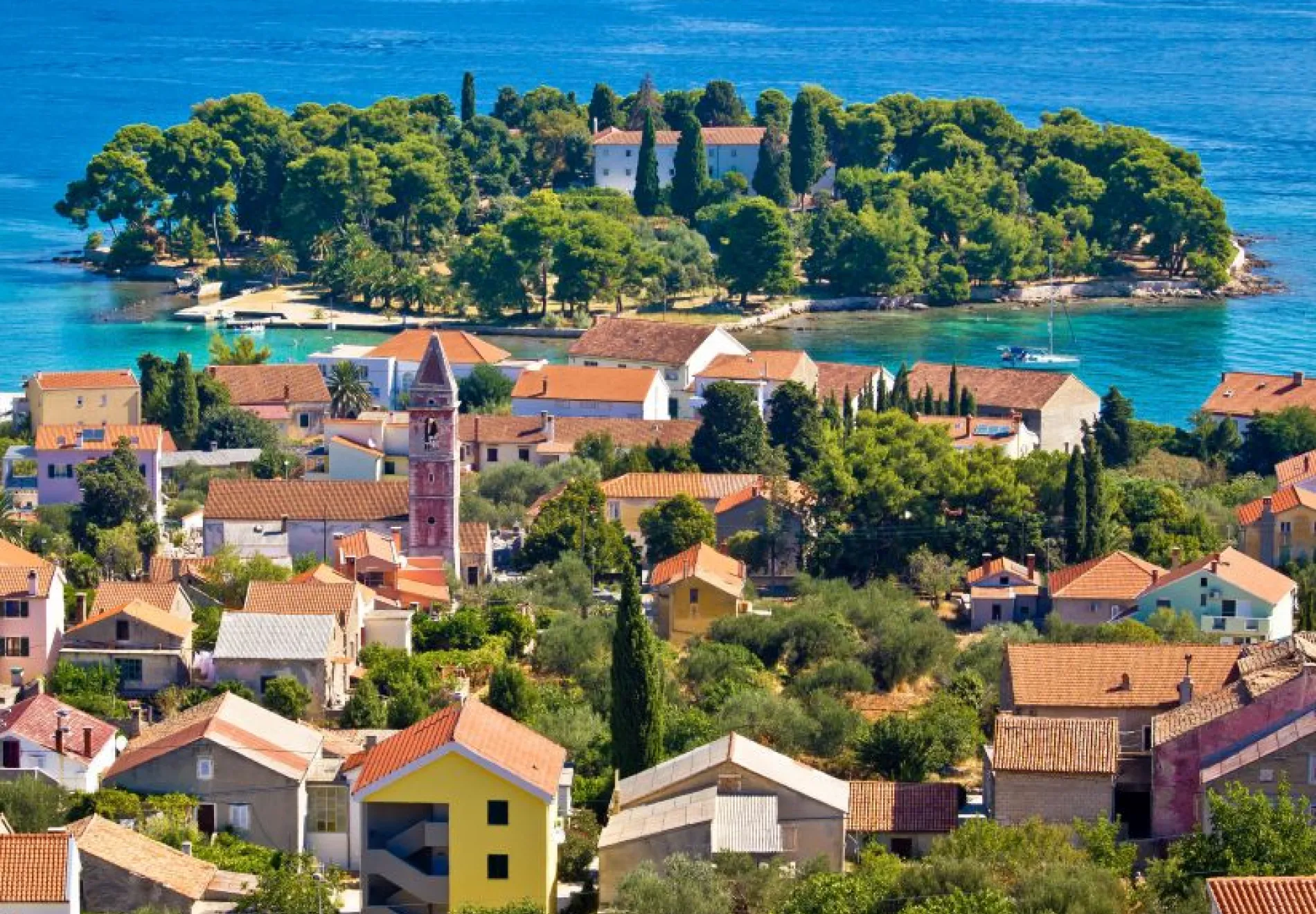Colorful houses with red-tiled roofs cluster near a church tower, with lush greenery and a small, tree-covered island beyond, all beside bright blue water under a clear sky.