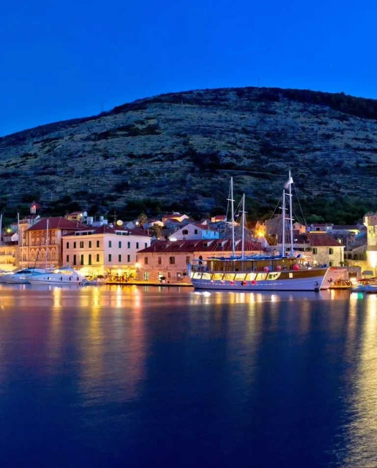 A peaceful coastal town at dusk with boats docked along the waterfront, illuminated buildings, and a hill rising in the background, all reflected in the calm water.