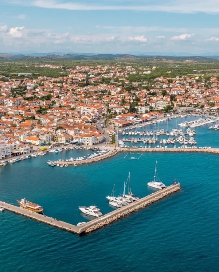 Aerial view of a coastal town with red-roofed buildings, a marina filled with boats, and turquoise water. A breakwater extends into the sea, sheltering the docked yachts and sailboats.