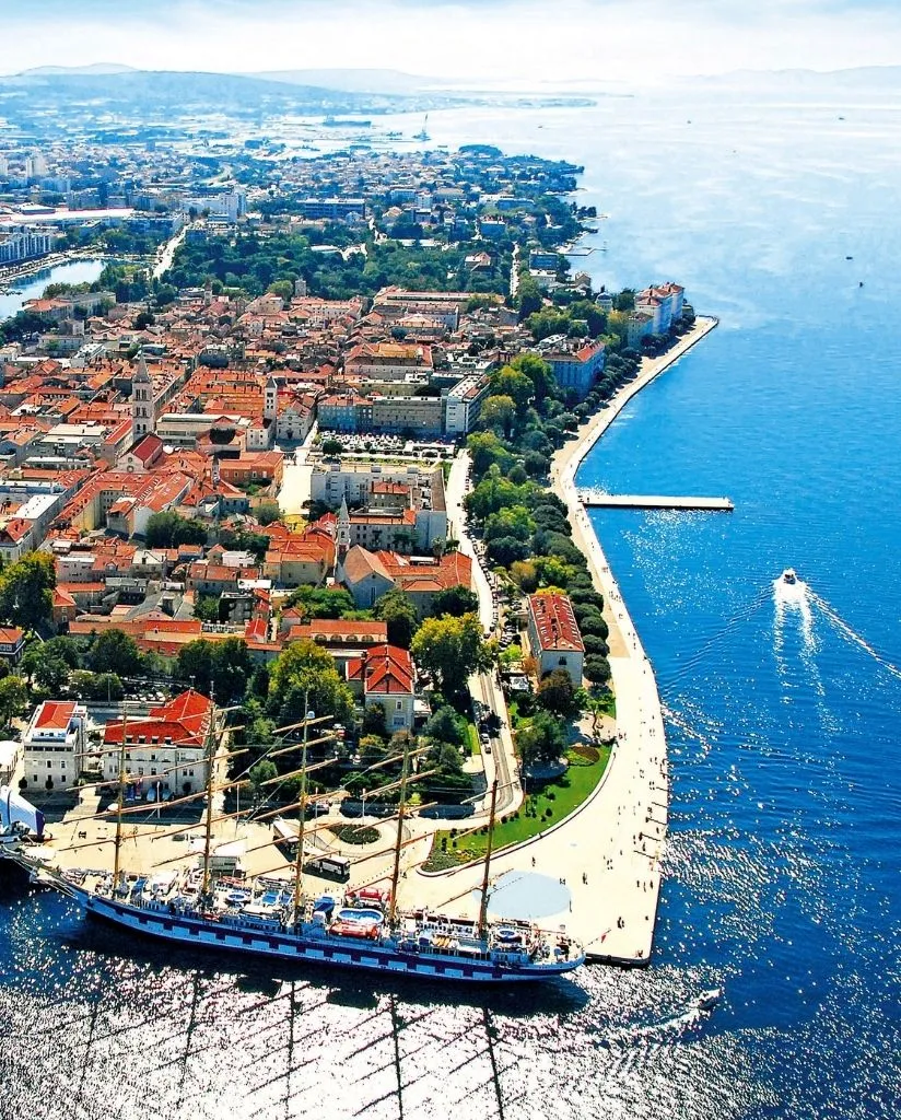 Aerial view of a coastal city with red-roofed buildings, a tree-lined promenade, a large sailing ship docked at the waterfront, and boats moving through bright blue seawater under a clear sky.
