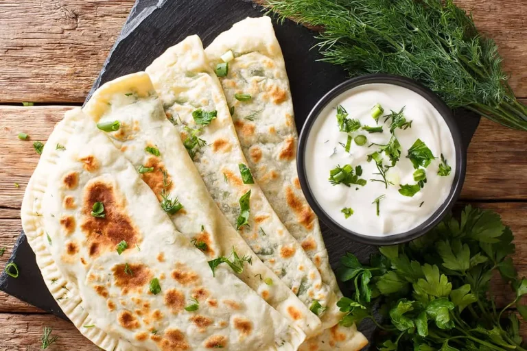 Three folded, golden-brown flatbreads with herbs are served on a slate board next to a bowl of white dipping sauce garnished with herbs, with fresh dill and parsley on the side, all on a rustic wooden surface.