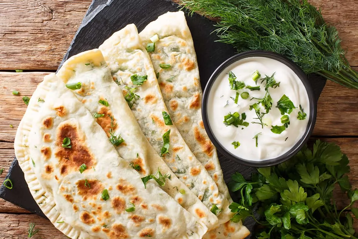 Three folded, golden-brown flatbreads with herbs are served on a slate board next to a bowl of white dipping sauce garnished with herbs, with fresh dill and parsley on the side, all on a rustic wooden surface.
