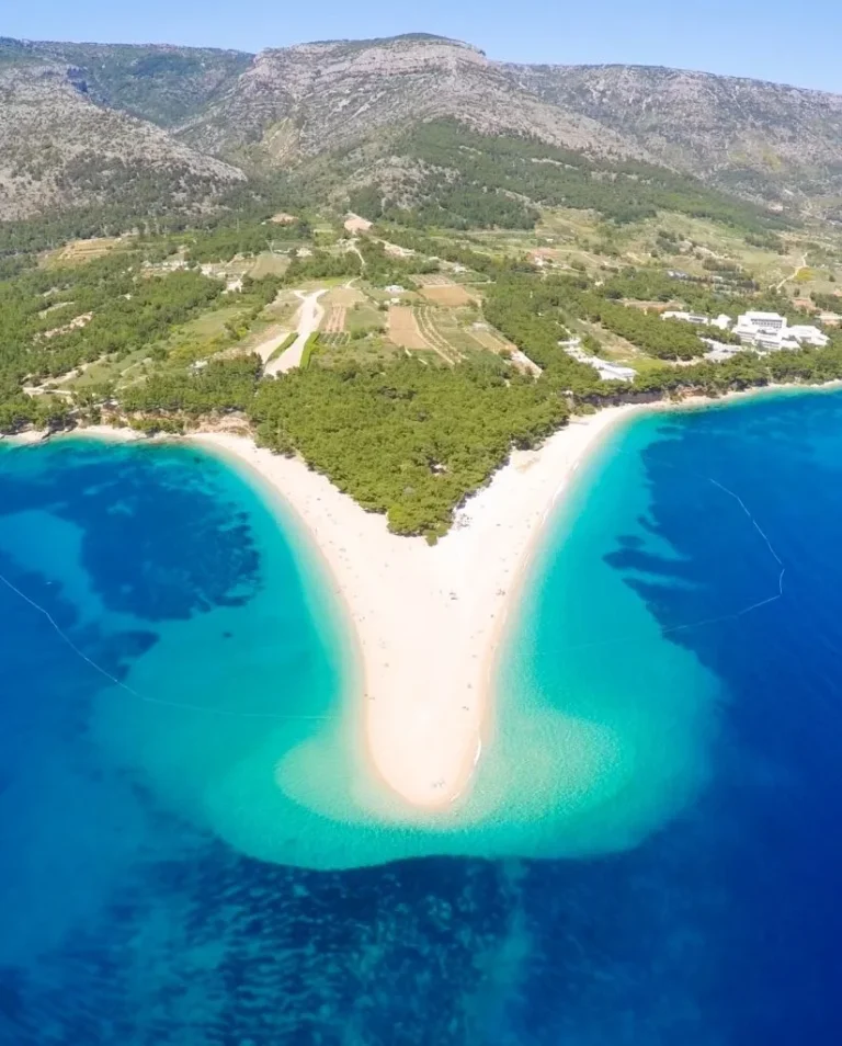 Aerial view of Zlatni Rat beach in Croatia, showing a narrow, sandy spit surrounded by clear turquoise water, with dense green trees and hills in the background.