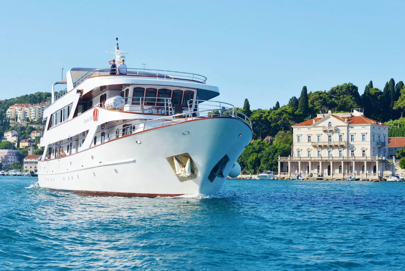 A large white yacht cruises on bright blue water near a shoreline with a grand building and lush green trees under a clear blue sky.