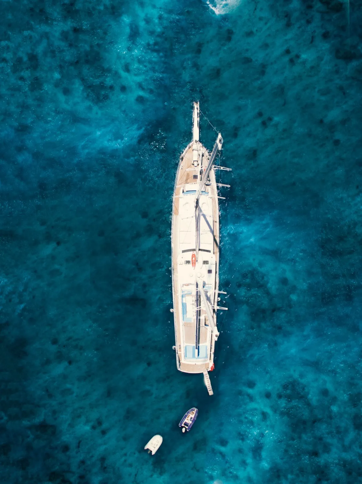 Aerial view of a large sailboat anchored in vibrant blue water with two small boats floating nearby. The sea below appears clear with varying shades of blue.