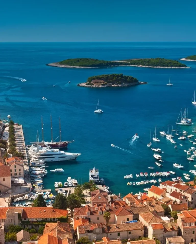 Aerial view of a coastal town with red-tiled rooftops, a marina filled with yachts and boats, and several small green islands surrounded by clear blue sea under a bright sky.