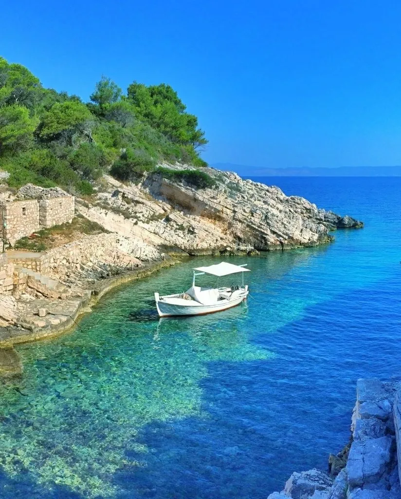 A small white boat with a canopy floats on clear turquoise water in a rocky cove, surrounded by stone walls and lush green trees under a bright blue sky.
