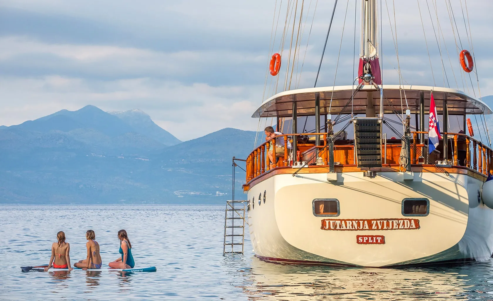 Three people sit on paddleboards near a large wooden boat named Jutarnja Zvijezda anchored on calm water, with mountains and a cloudy sky in the background.