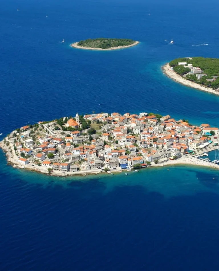 Aerial view of a small, densely built island town surrounded by deep blue sea, with orange-roofed buildings, a church, marinas, and two nearby green islets.