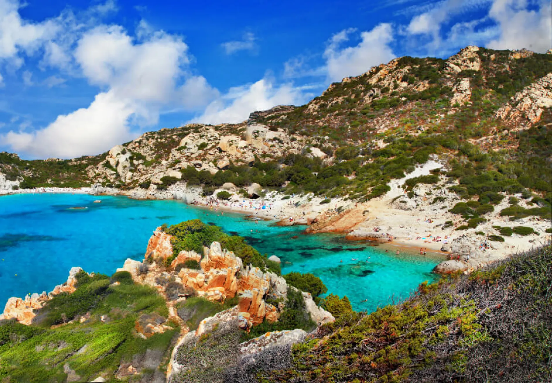 A stunning coastal landscape with turquoise blue water, a sandy beach, rocky cliffs, green vegetation, and partly cloudy skies. People are visible relaxing on the beach and swimming in the clear sea.