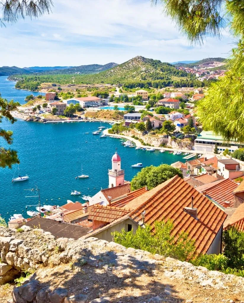 A coastal town with red-roofed buildings, a church with a pink tower, and boats docked along a blue bay, surrounded by green hills and trees under a clear sky.
