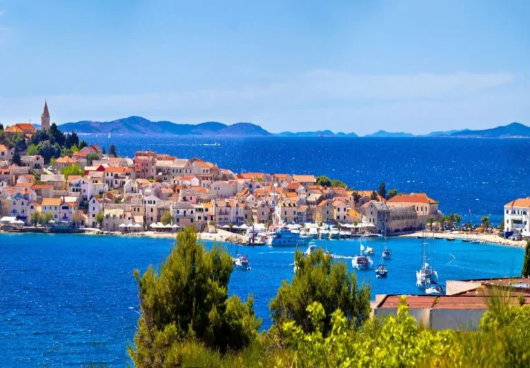 A coastal town with red-roofed buildings sits by the blue sea, dotted with boats and surrounded by green trees, with distant hills and islands under a clear, sunny sky.