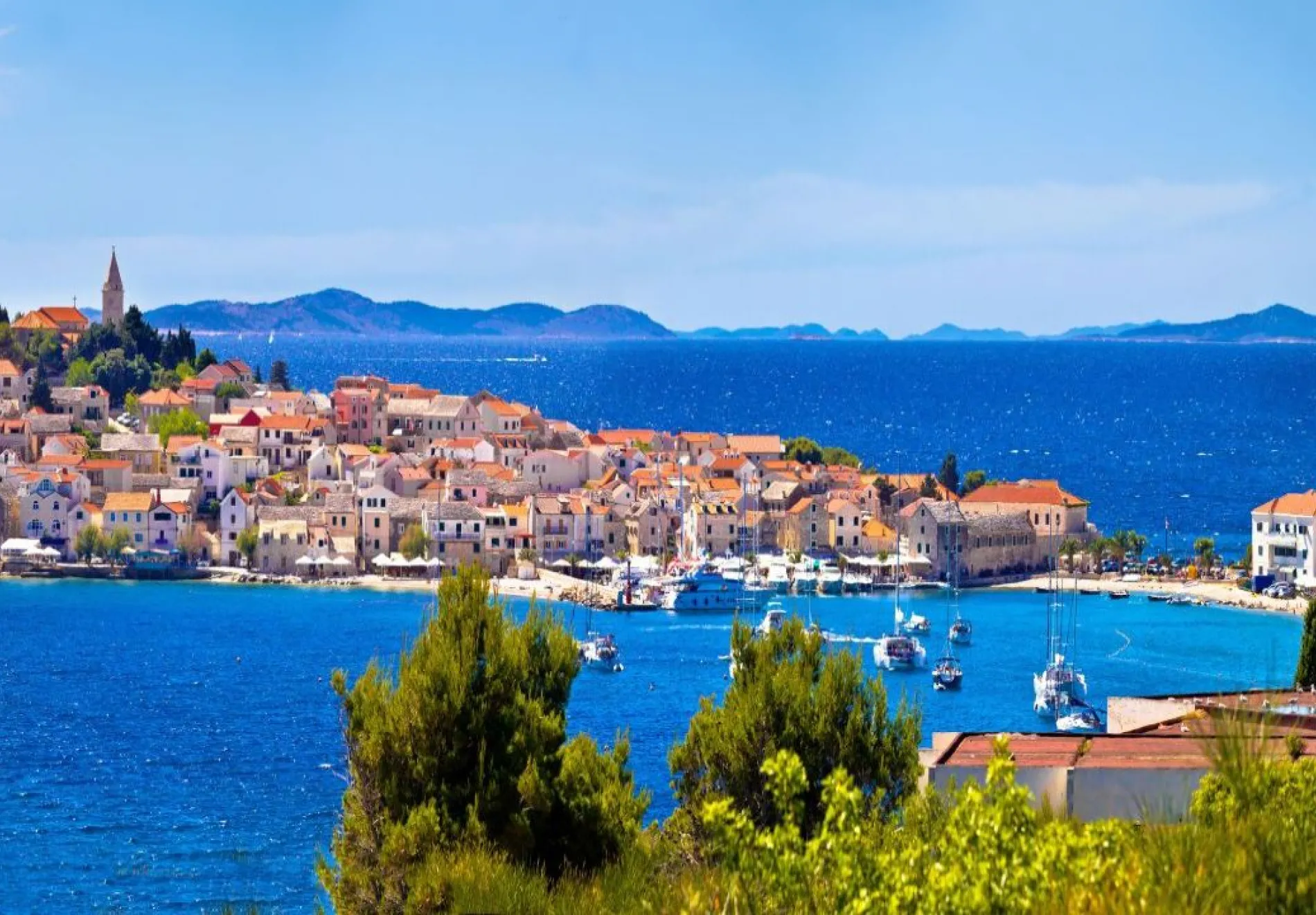 A coastal town with red-roofed buildings sits by the blue sea, dotted with boats and surrounded by green trees, with distant hills and islands under a clear, sunny sky.
