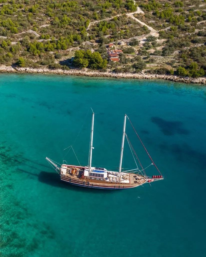 Aerial view of a sailboat with two masts anchored in clear turquoise water near a rocky, tree-lined coastline with a small house visible among the greenery.