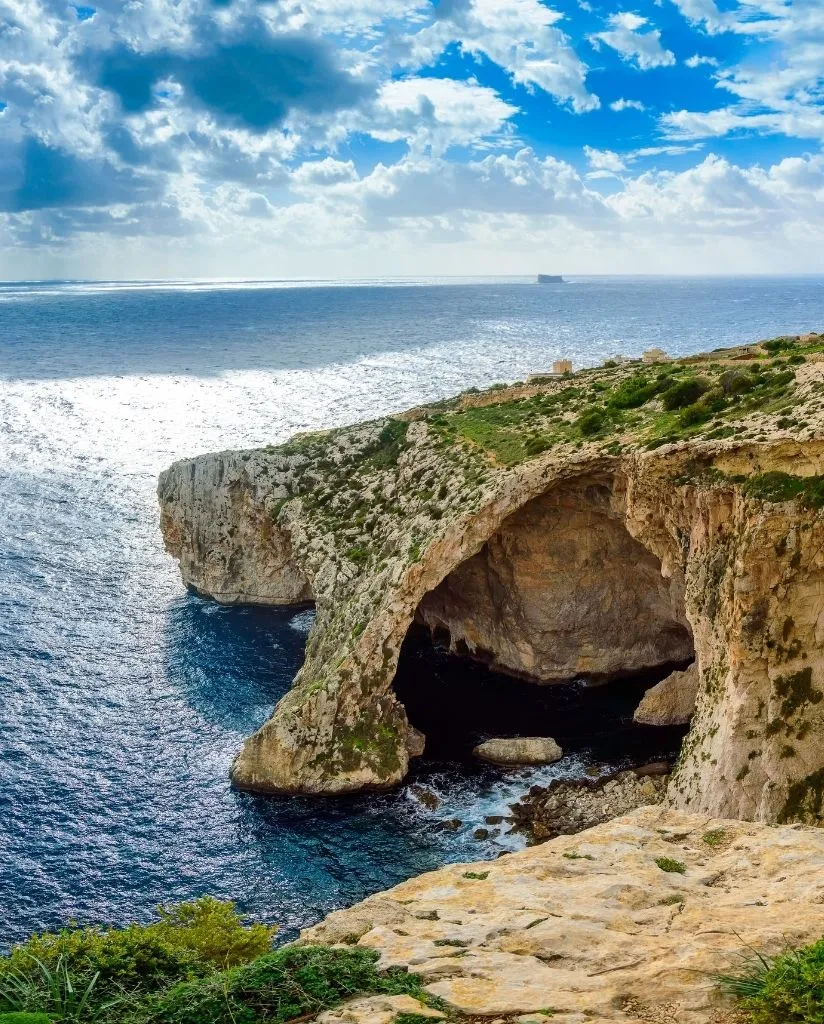 A natural rock arch juts out over deep blue sea water, with sunlight reflecting off the surface. The scene is set under a partly cloudy sky, and green vegetation grows on the rocky cliff.