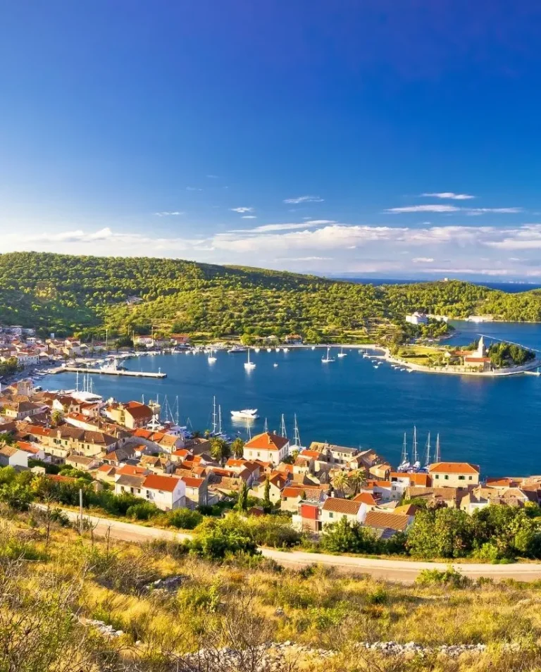 A panoramic view of a coastal village with red-roofed houses, a harbor filled with boats, green hills, and a bright blue sky with scattered clouds.