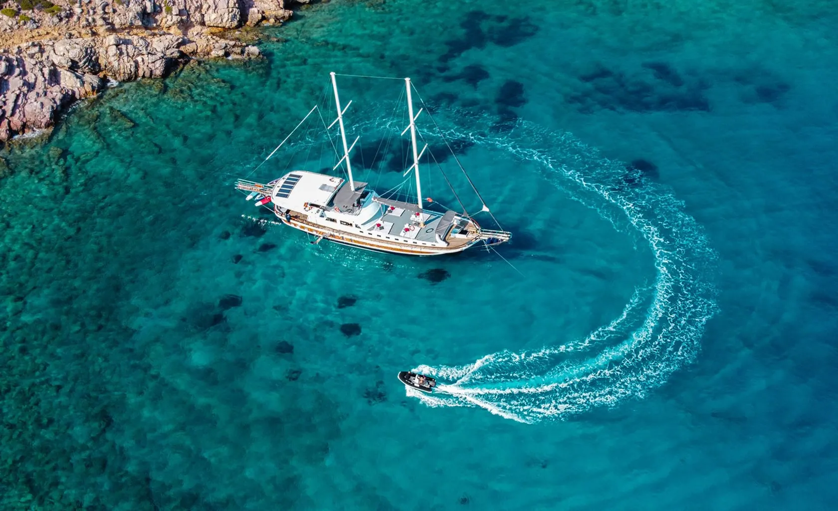 Aerial view of a large white sailboat anchored in clear turquoise water near a rocky shoreline, as a small motorboat creates a circular wake nearby.