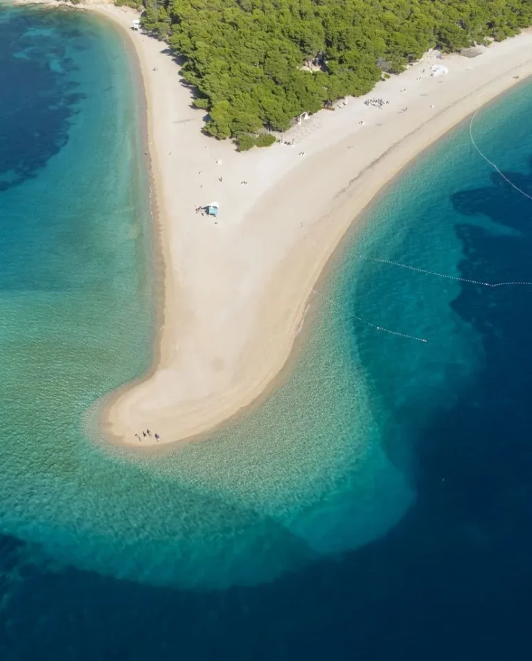 Aerial view of a sandy beach peninsula extending into clear blue sea, bordered by turquoise and deep blue water, with green forest at the shore and a few people visible on the sand.