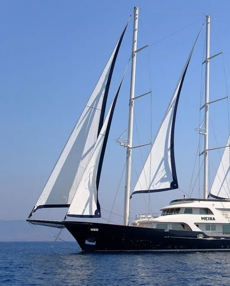 A large, luxurious sailboat with three tall masts and white sails glides on calm blue water under a clear sky. The boats name, MEIRA, is visible on the side.
