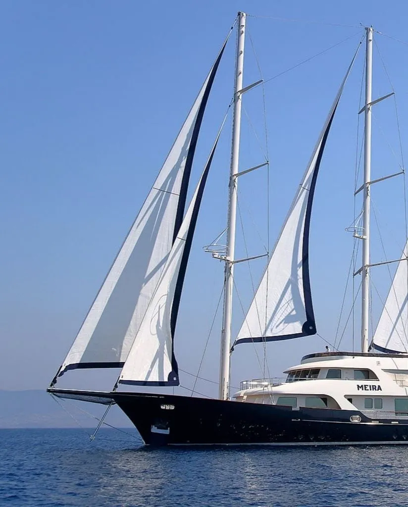 A large, luxurious sailboat with three tall masts and white sails glides on calm blue water under a clear sky. The boats name, MEIRA, is visible on the side.