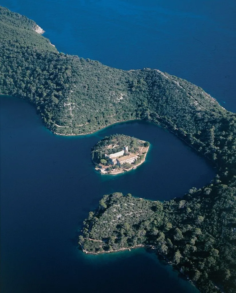 A small, forested island with a large, rectangular building sits in a blue lake surrounded by wooded hills, creating a striking contrast between water and greenery.