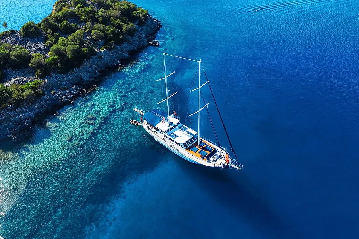 Aerial view of a white sailboat anchored near a rocky, tree-covered island, surrounded by clear turquoise and deep blue water.
