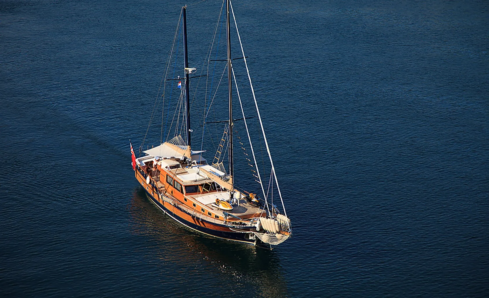 A wooden sailboat with two masts and a spacious deck is sailing on calm, deep blue water. The boat has white sails furled and a red flag at the stern.