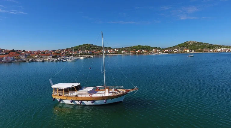 A wooden sailboat floats on calm blue water near a coastal town, with hills and houses visible under a clear blue sky in the background.