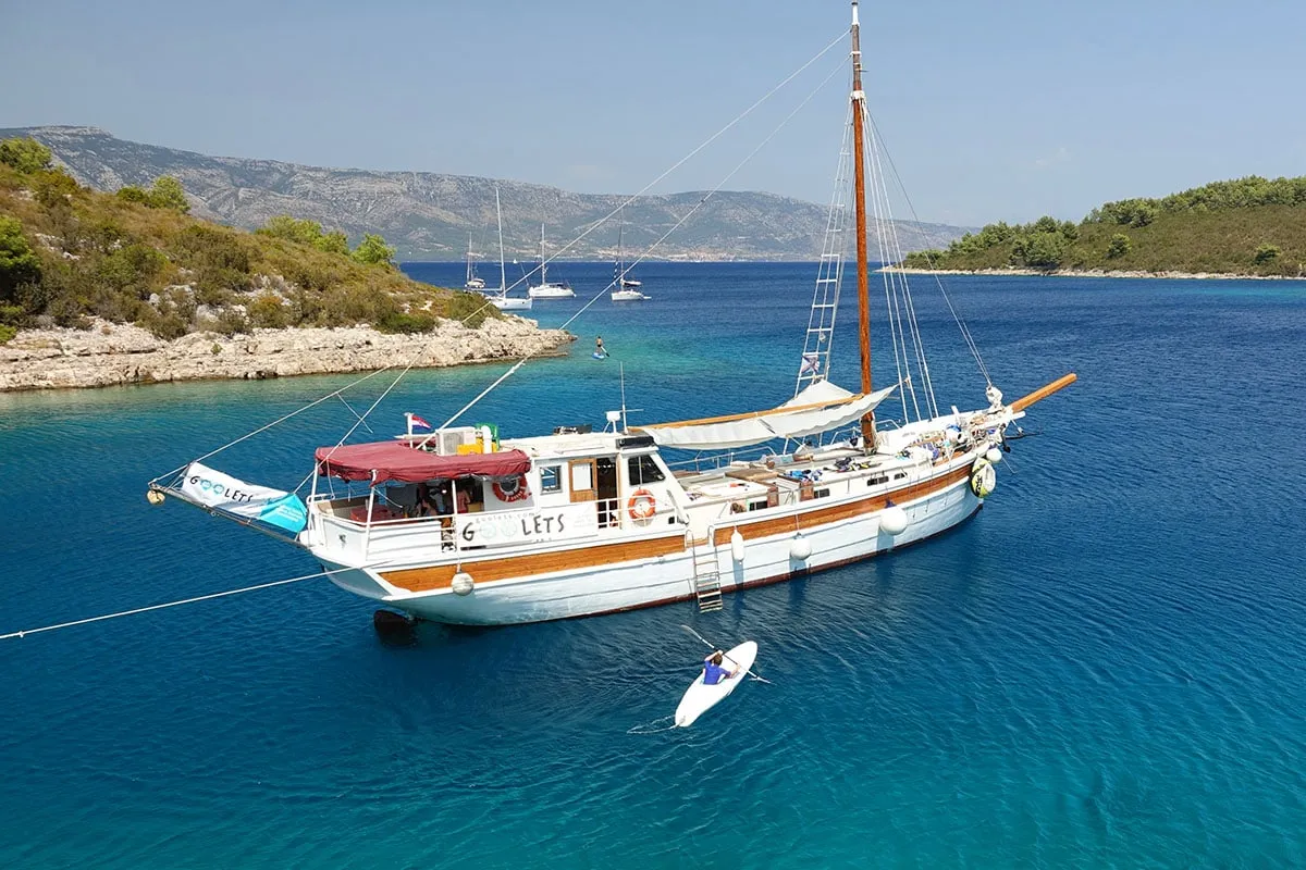 A wooden sailboat anchored in clear blue water near a rocky, green coastline. Several sailboats are in the distance and a person paddles a small white kayak beside the boat under a sunny sky.