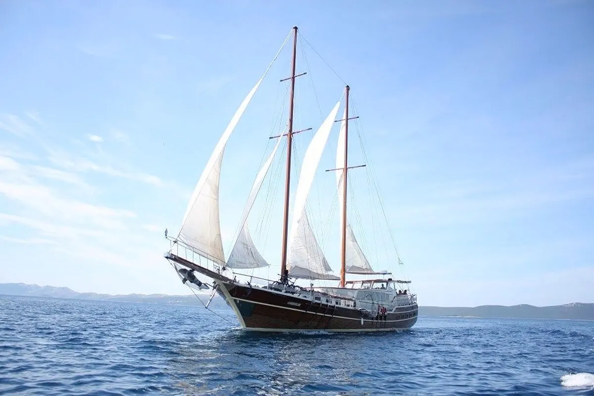 A large wooden sailboat with white sails unfurled glides across calm blue water under a clear sky, with distant land visible on the horizon.