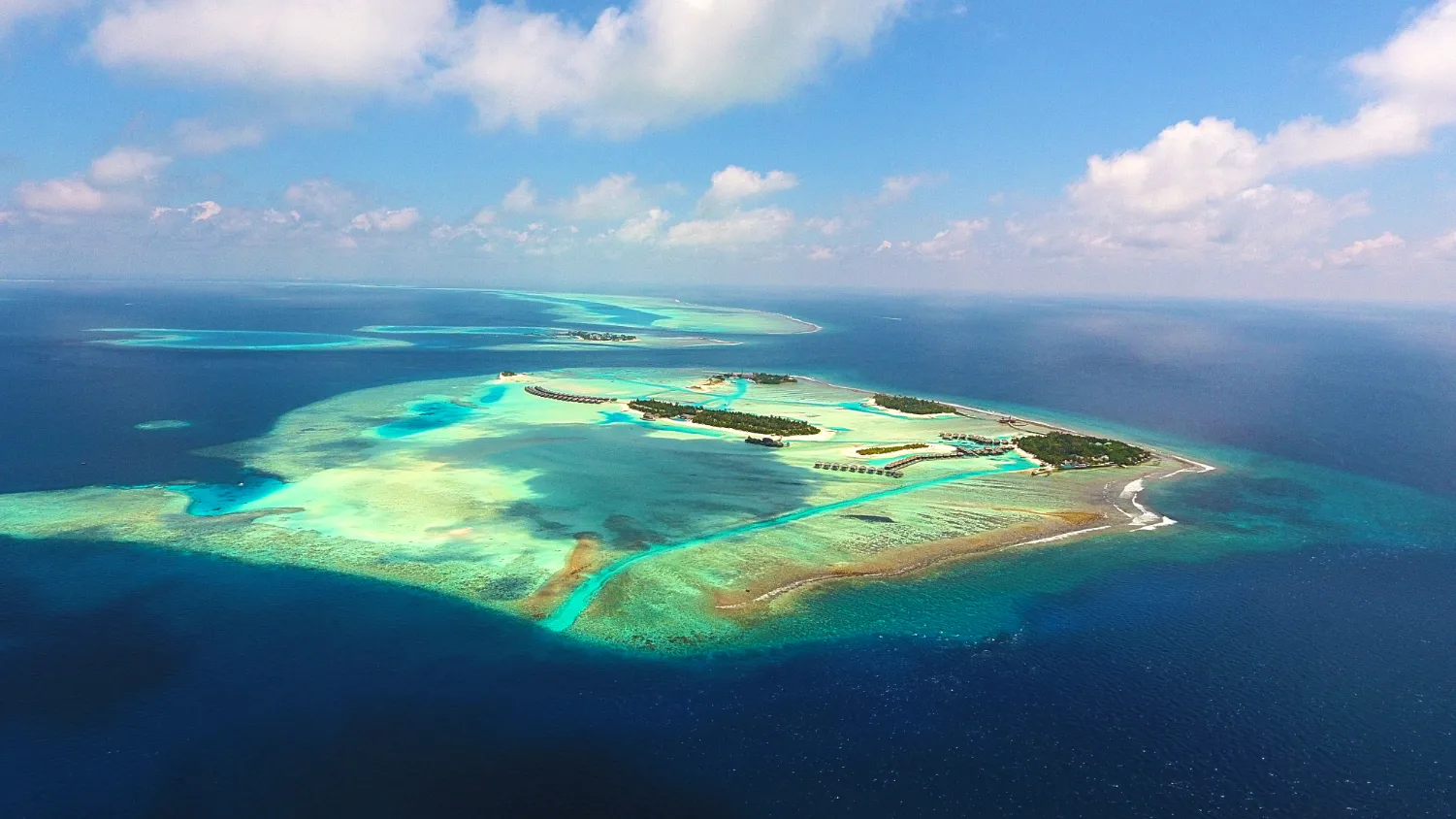 Aerial view of a tropical island with turquoise shallow waters, sandy beaches, and lush greenery, surrounded by deep blue ocean under a partly cloudy sky.