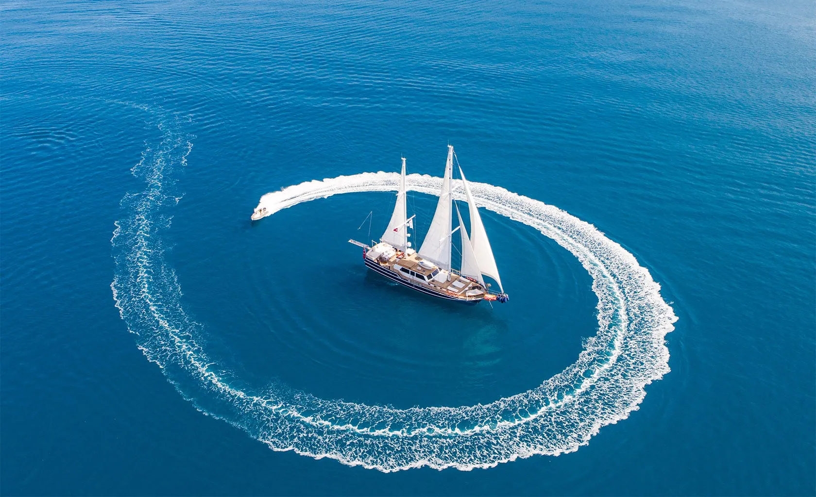Aerial view of a sailboat on calm blue water, with a small motorboat creating a circular white trail around it. The scene emphasizes the contrast between the still sailboat and the dynamic motion of the motorboat.
