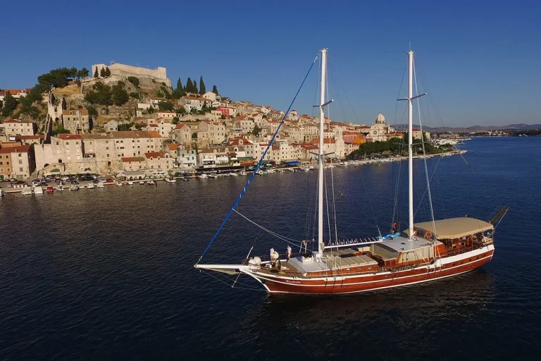 A large wooden sailboat cruises on calm blue water near a coastal town with terracotta-roofed buildings and a hilltop fortress under a clear blue sky.