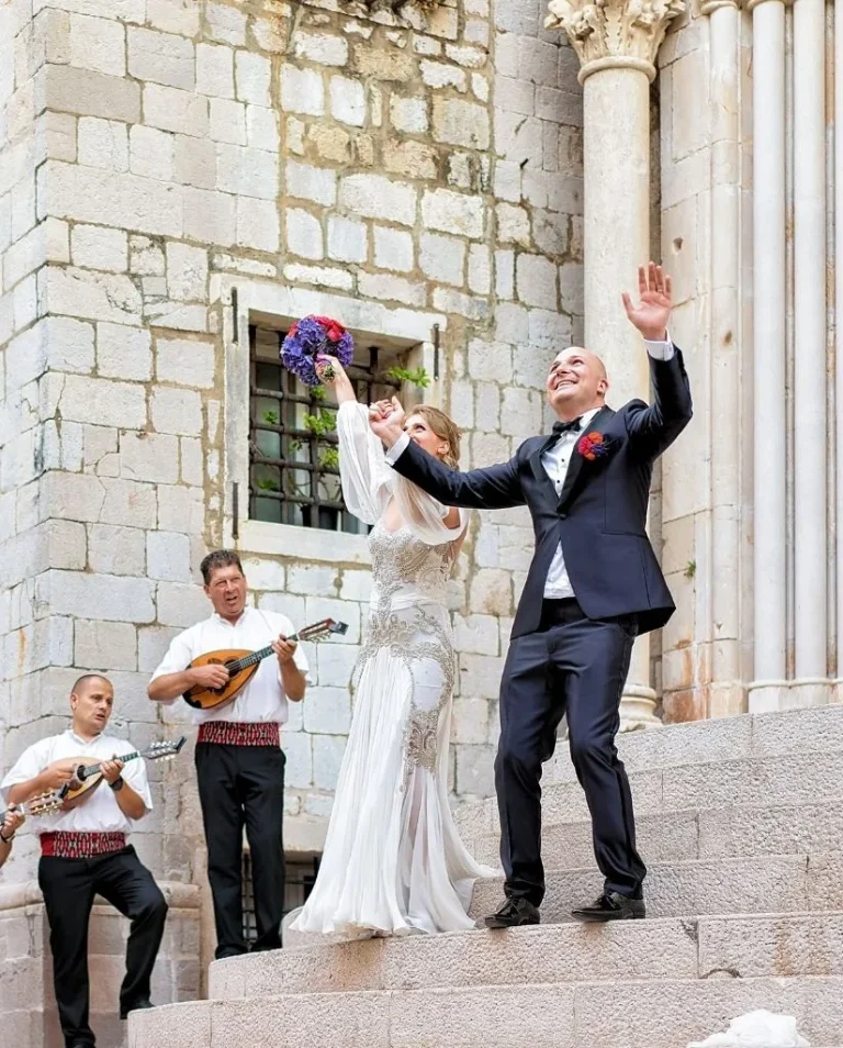 A bride and groom joyfully celebrate on stone steps, with the bride holding a bouquet in the air. Two musicians in white shirts play string instruments nearby against a historic stone building backdrop.
