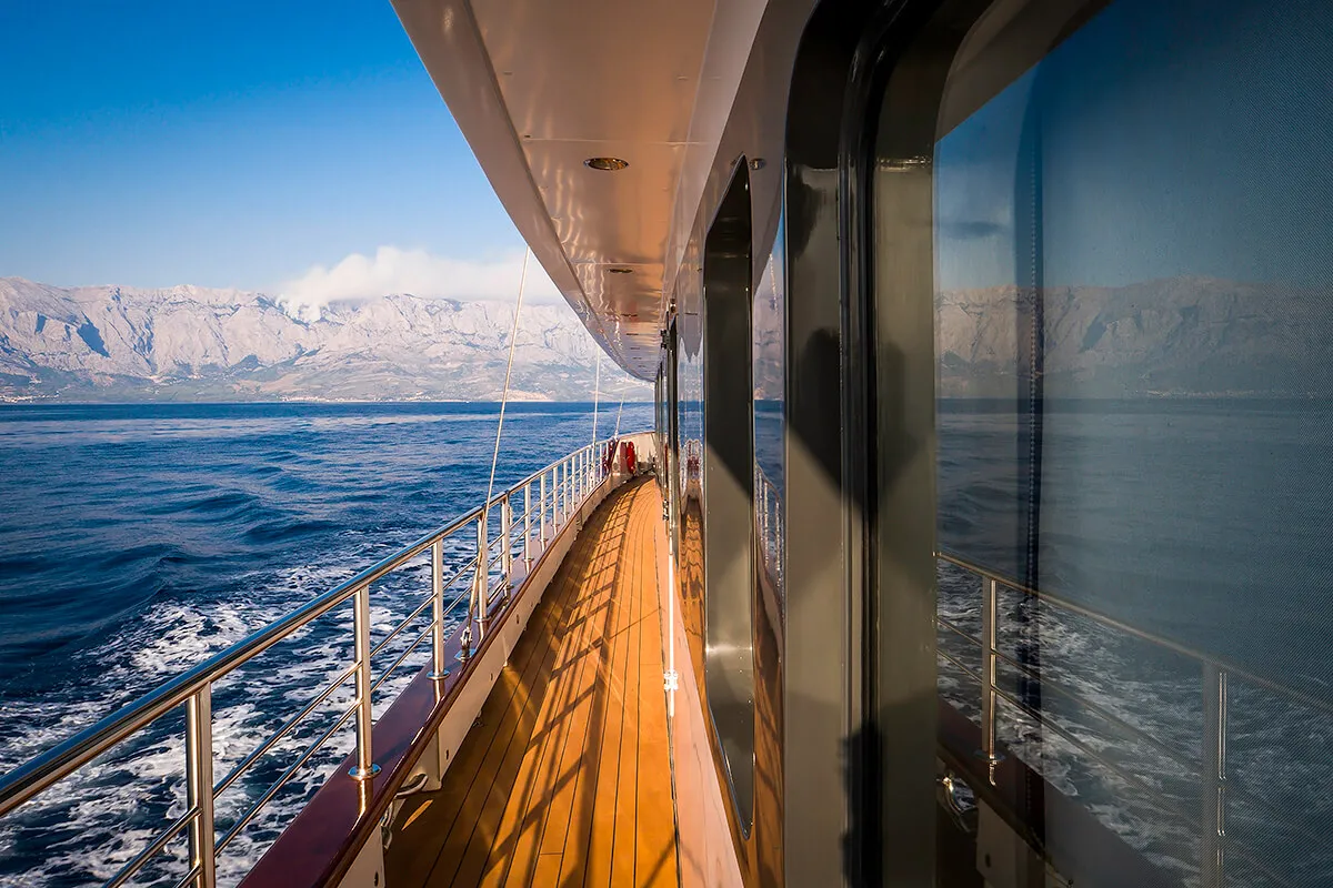 A view from the deck of a boat sailing on calm blue water, with mountains in the background and reflections of the scene in the boats windows under a clear sky.