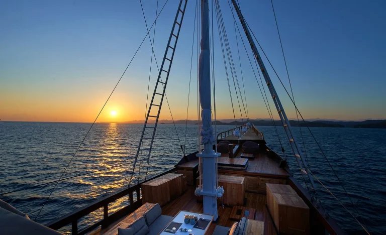 A view from the deck of a wooden sailboat at sunset, with the sun low over the horizon, calm sea waters, and distant land visible under a clear sky.