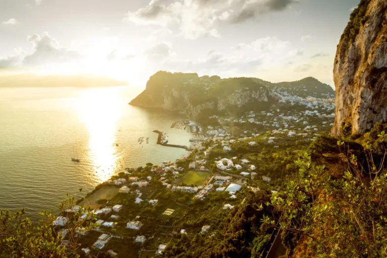 A scenic view of the island of Capri, Italy, at sunset shows sparkling waters, dramatic cliffs, and a town with white buildings nestled along the coastline. Lush greenery covers much of the landscape.