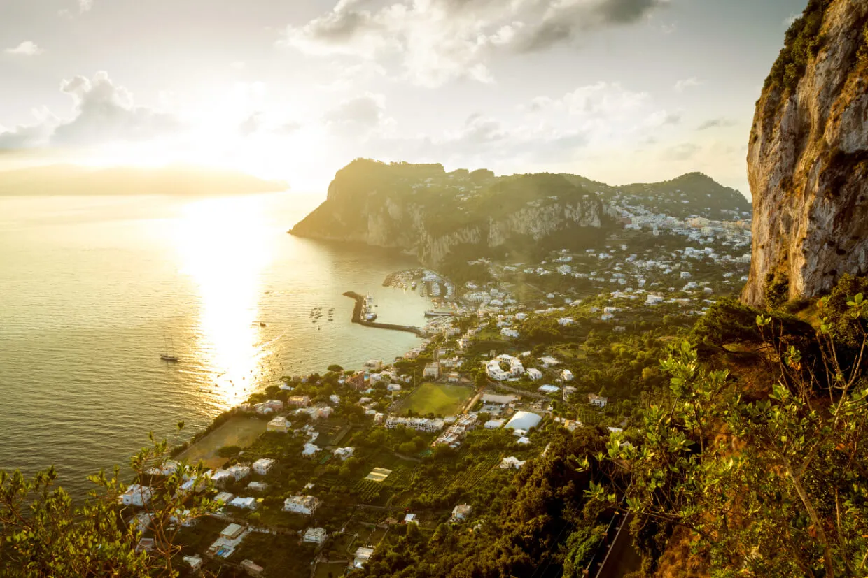 A scenic view of the island of Capri, Italy, at sunset shows sparkling waters, dramatic cliffs, and a town with white buildings nestled along the coastline. Lush greenery covers much of the landscape.