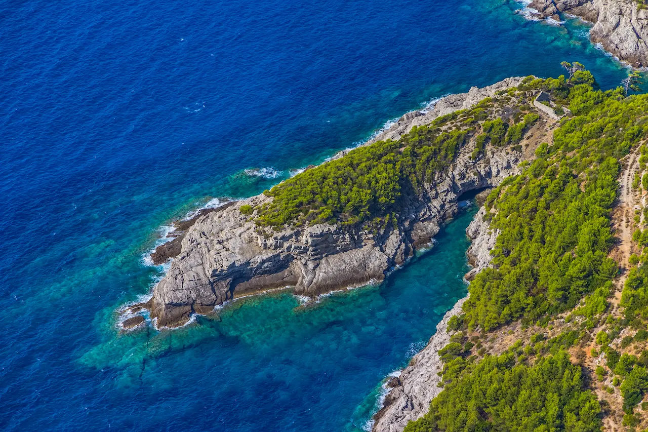 A rocky peninsula covered with green trees juts into clear, vibrant blue ocean water, creating a striking contrast between land and sea. The coastline features rugged rocks and lush vegetation.