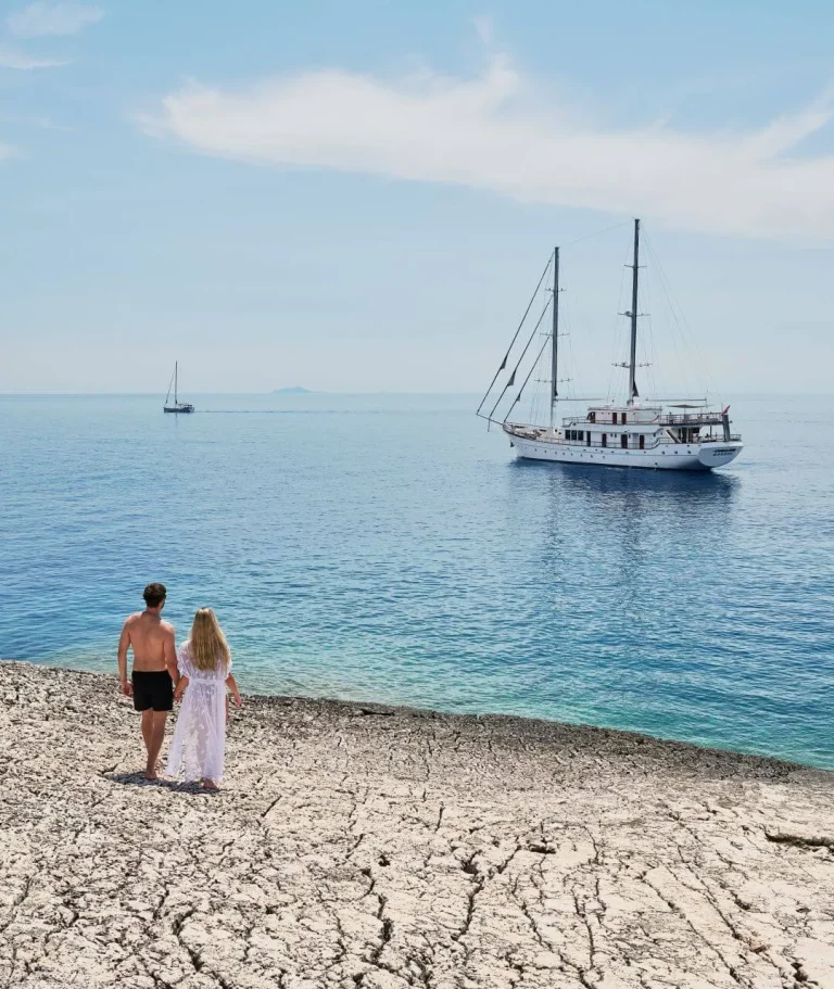 A man and woman stand on a rocky shore, holding hands and looking out at a large sailboat anchored in calm blue water, with another sailboat visible in the distance under a clear sky.