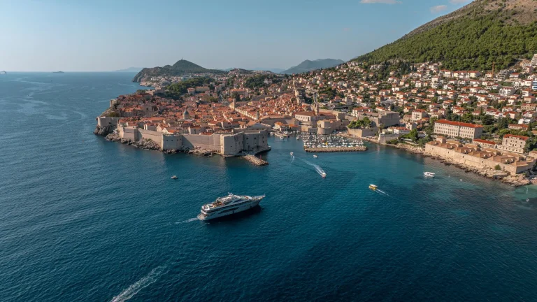 Aerial view of Dubrovnik, Croatia, showing the historic walled old town by the sea, surrounded by hills and modern houses, with several boats sailing on the blue water.