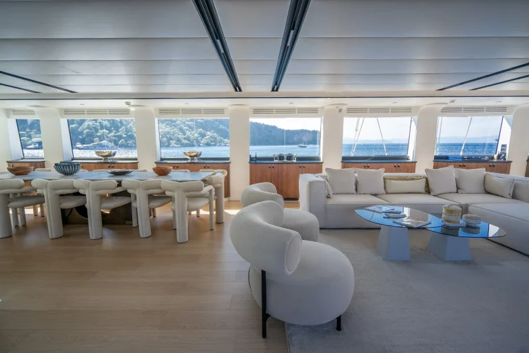 Spacious yacht interior with large windows, white modern furniture, a dining table with chairs, and a cozy seating area with sofas and a glass coffee table, overlooking the ocean and distant shoreline.