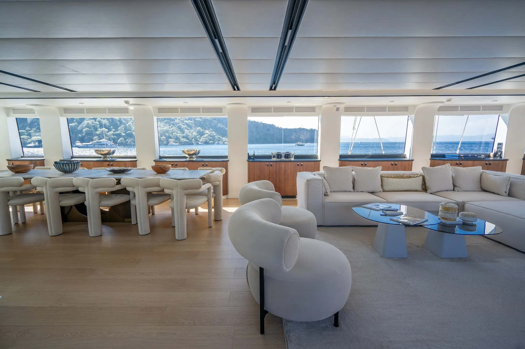 Spacious yacht interior with large windows, white modern furniture, a dining table with chairs, and a cozy seating area with sofas and a glass coffee table, overlooking the ocean and distant shoreline.