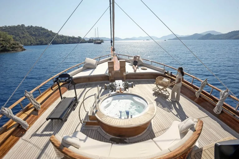 A woman stands beside a round jacuzzi on the deck of a luxury yacht, surrounded by lounge chairs and exercise equipment, with blue sea and distant green hills under a clear sky.