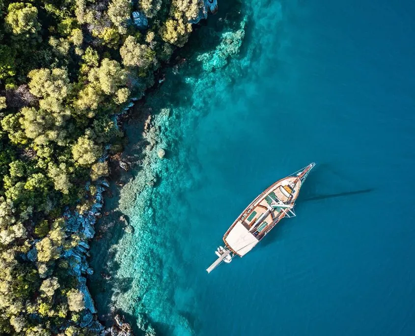 Aerial view of a wooden sailboat floating on clear blue water near a lush, green, tree-lined coastline with rocks visible under the water near the shore.
