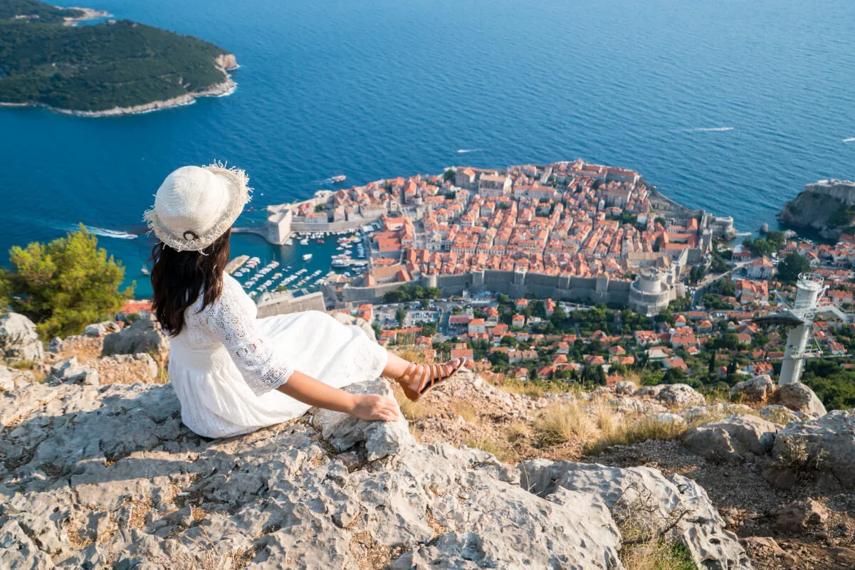 A woman in a white dress and hat sits on a rocky hilltop, overlooking the red-roofed buildings and historic walls of Dubrovnik, Croatia, with the blue sea in the background.