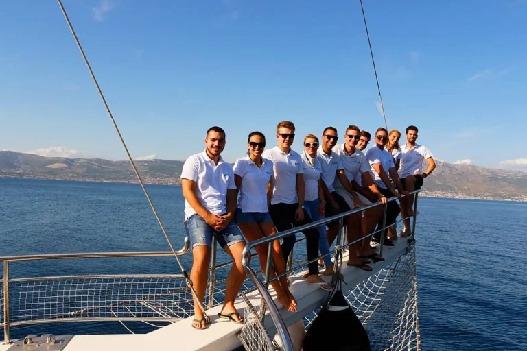 A group of people in white shirts and sunglasses stand together on the bow of a sailboat, smiling, with calm blue sea and distant coastline in the background.